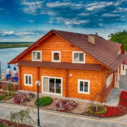 Drewniany dom z brązowym dachem, położony nad jeziorem, z brukowanym podjazdem i rabatami kwiatowymi.