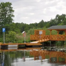Drewniany pomost z zadaszeniem i znakiem parkingu nad wodą, otoczony trzciną i drzewami.