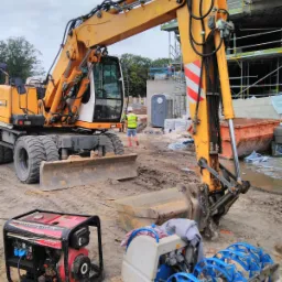 Ż&oacute;łta koparka kołowa na placu budowy z widocznym agregatem prądotw&oacute;rczym, zgrzewarką do rur na palecie, kontenerem, przenośną toaletą i fragmentem budynku w rusztowaniach w tle.