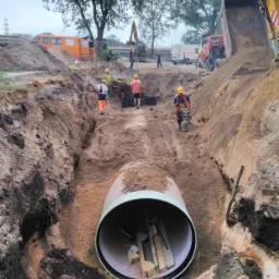 Montaż rury kanalizacyjnej w wykopie, widoczni pracownicy w kaskach i kamizelkach odblaskowych, zagęszczarka, ciężarówka wywrotka w tle.