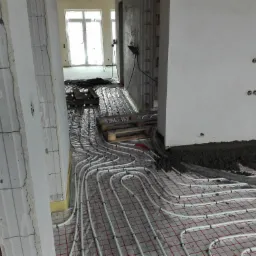 Montaż ogrzewania podłogowego: białe rurki rozłożone na folii z czerwoną siatką, częściowo zalane jastrychem, w tle palety i okna.