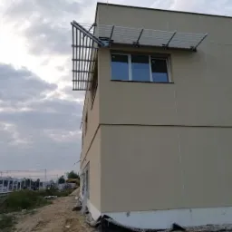 Nowoczesny budynek otynkowany na beżowo z metalową konstrukcją nad oknami, widok z zewnątrz na plac budowy z elementami konstrukcji stalowych w tle.