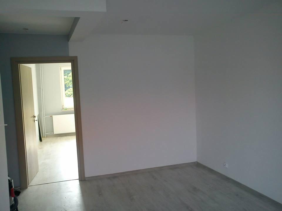 Wnętrze pokoju po malowaniu na biało, widoczne szare panele podłogowe, drzwi z klamką prowadzące do kolejnego pomieszczenia z oknem i grzejnikiem.