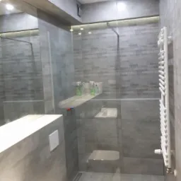 Nowoczesna łazienka wykończona szarymi płytkami z motywem miast, kabina prysznicowa typu walk-in z przezroczystą szybą, biała drabinka grzewcza, toaleta w minimalistycznym stylu.