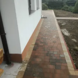 Nowa ścieżka z kostki brukowej w odcieniach brązu i beżu, prowadząca wzdłuż białej ściany domu z widocznymi oknami i rynną, obok rabata z przygotowaną ziemią pod nasadzenia.