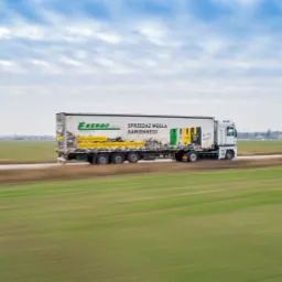 Ciężarówka z naczepą marki Energo z napisem 'Sprzedaż węgla kamiennego' w ruchu na drodze, widok z boku na tle zielonego pola i pochmurnego nieba.