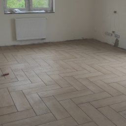 HomeDesign - Ułożona podłoga z płytek imitujących drewno w jodełkę francuską w jasnym, surowym wnętrzu z grzejnikiem, oknem i wyjściem na balkon.