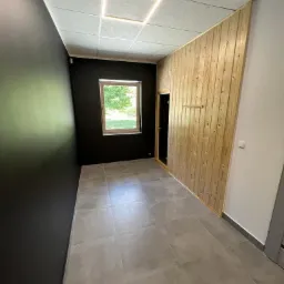 Wnętrze pomieszczenia z szarą podłogą z płytek, czarną ścianą, oknem i ścianą wykończoną jasnym drewnem z ukrytymi drzwiami, oświetlenie sufitowe w kształcie kwadratu.