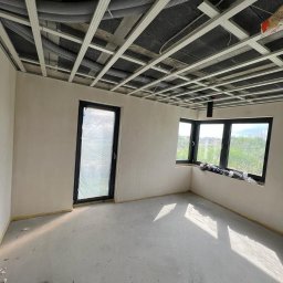 HomeDesign - Surowy stan wnętrza z widoczną konstrukcją sufitu podwieszanego z szarego bloku betonowego, rurami wentylacyjnymi i aluminiowym stelażem, ściany przygotowane do malowania, okna z czarnymi ramami.
