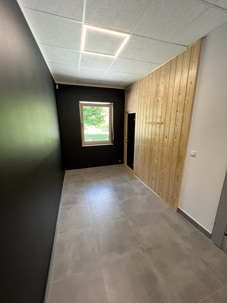 Wnętrze pomieszczenia z szarą podłogą z płytek, czarną ścianą, oknem i ścianą wykończoną jasnym drewnem z ukrytymi drzwiami, oświetlenie sufitowe w kształcie kwadratu.