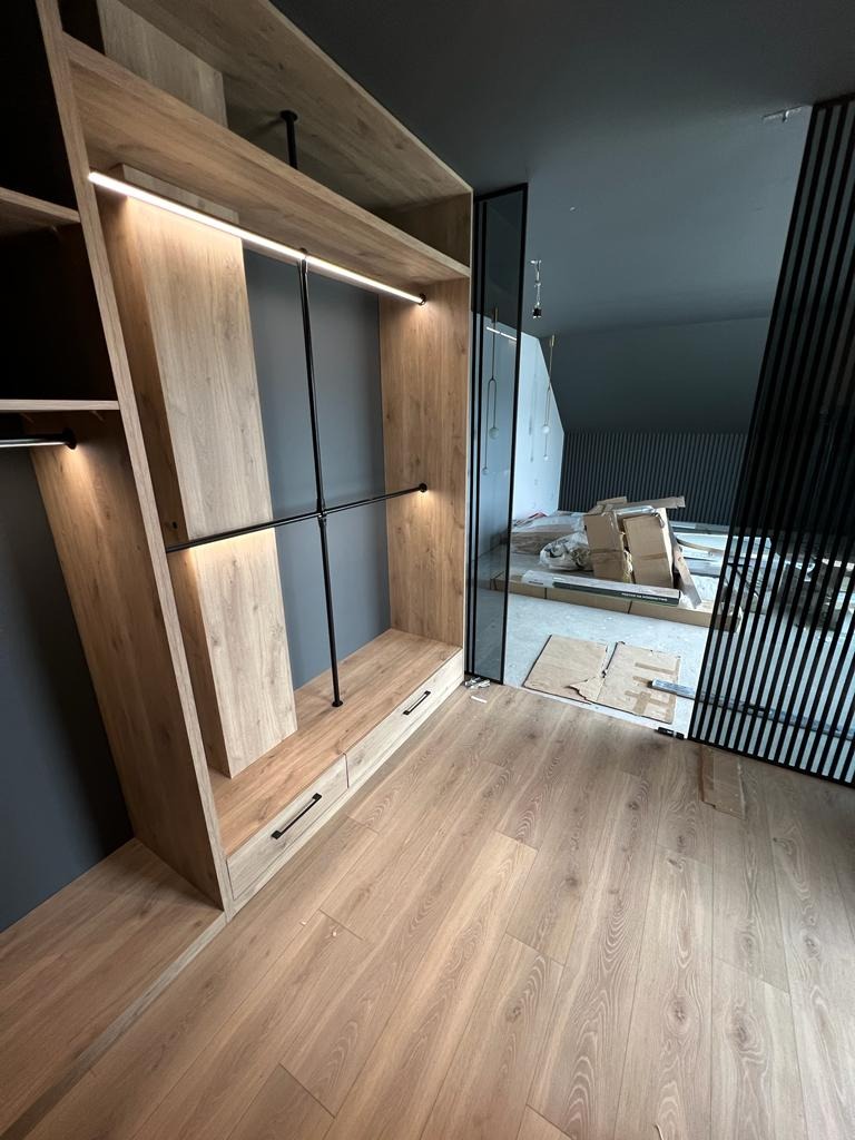 Jasny, drewniany system szaf wnękowych z oświetleniem LED, czarnymi relingami i szufladami, na podłodze panele w odcieniach beżu i brązu, w tle widoczne kartony i elementy konstrukcyjne...