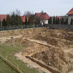 Wykopane rowy pod fundamenty domu jednorodzinnego, widoczny niwelator laserowy na statywie. W tle domy jednorodzinne i ogrodzenie.