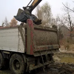 Koparka ładuje błotnistą ziemię na wywrotkę na tle wykopu. Widoczne drzewa i pochmurne niebo. Elementy maszyny intensywnie zabrudzone.