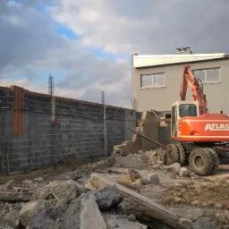 Pomarańczowy koparko-ładowarka Atlas na tle częściowo zburzonej ściany z szarej cegły i budynku. W tle pochmurne niebo.