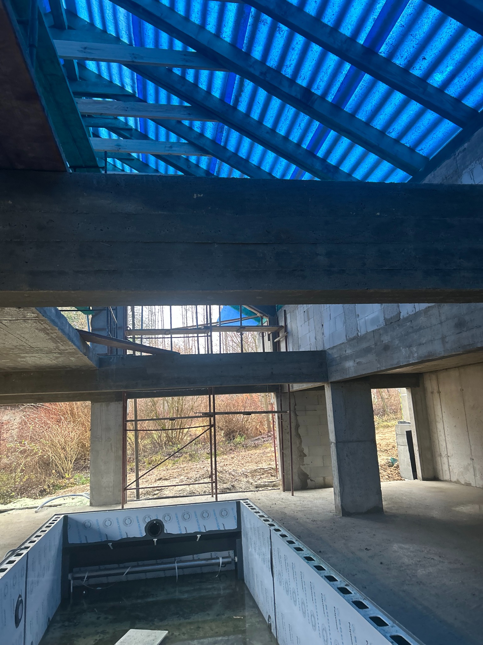 Surowy stan budynku w trakcie budowy: betonowe stropy, kolumny, ściany z pustaków, niebieskie zadaszenie i przygotowany basen w ziemi. Widok z wnętrza na zewnątrz.