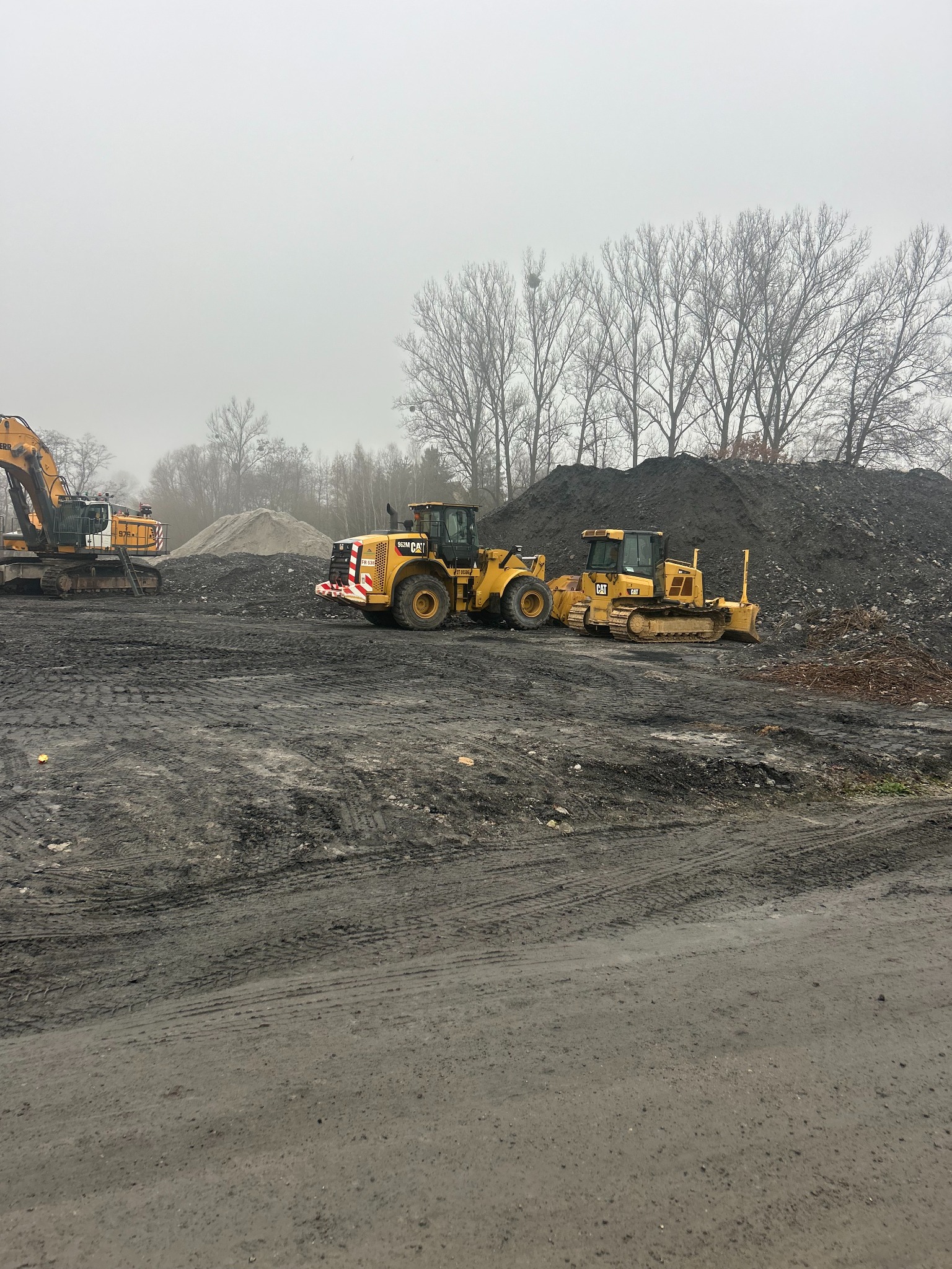 Żółte maszyny budowlane CAT (ładowarka i spycharka) na placu budowy z hałdami ziemi w tle, w pochmurny dzień. Widoczne ślady opon na ziemi.