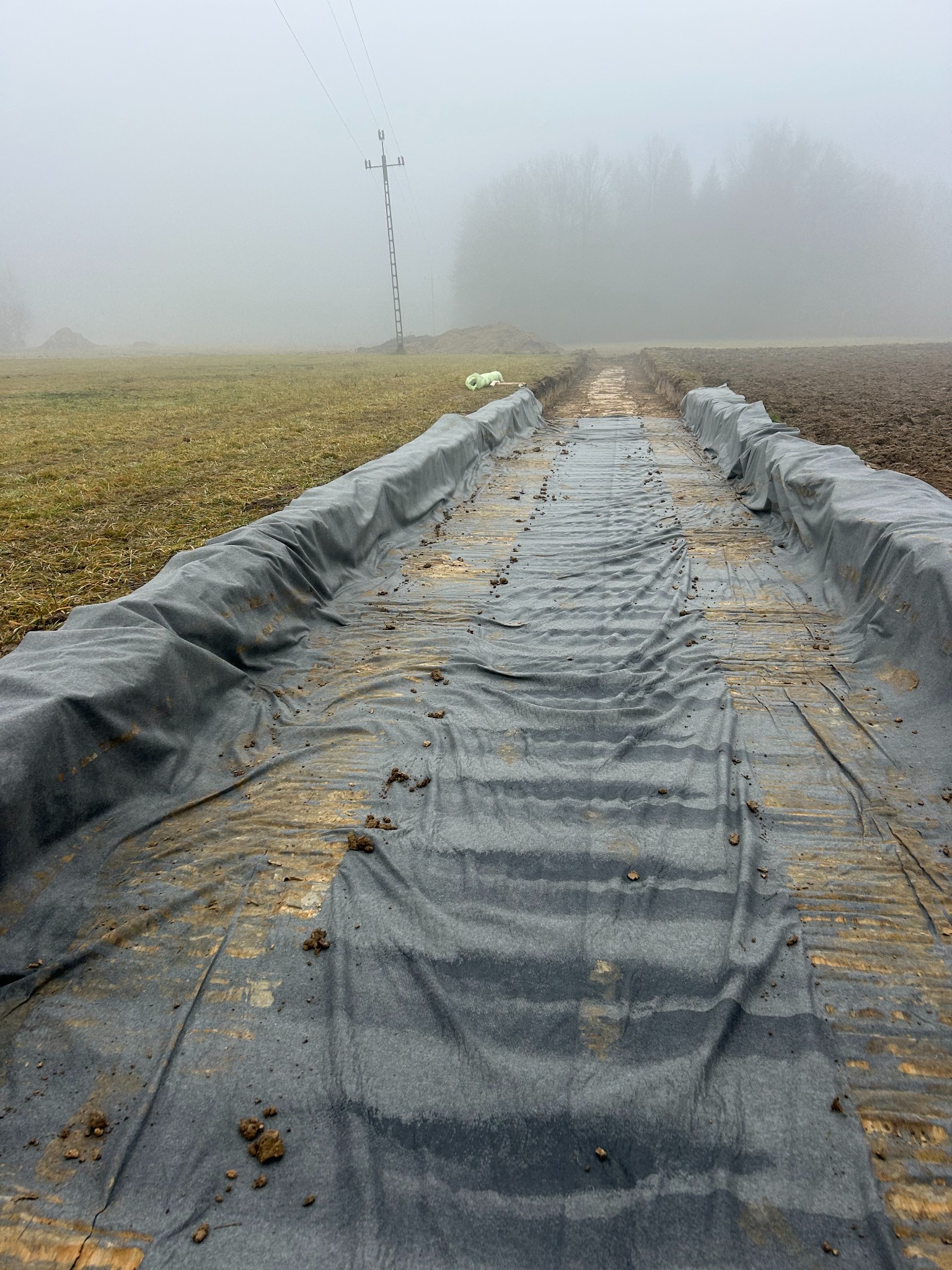 Przygotowanie terenu pod budowę drogi w mglisty dzień. Wykop wyłożony szarą geowłókniną, w tle słup wysokiego napięcia i zalesiony teren.