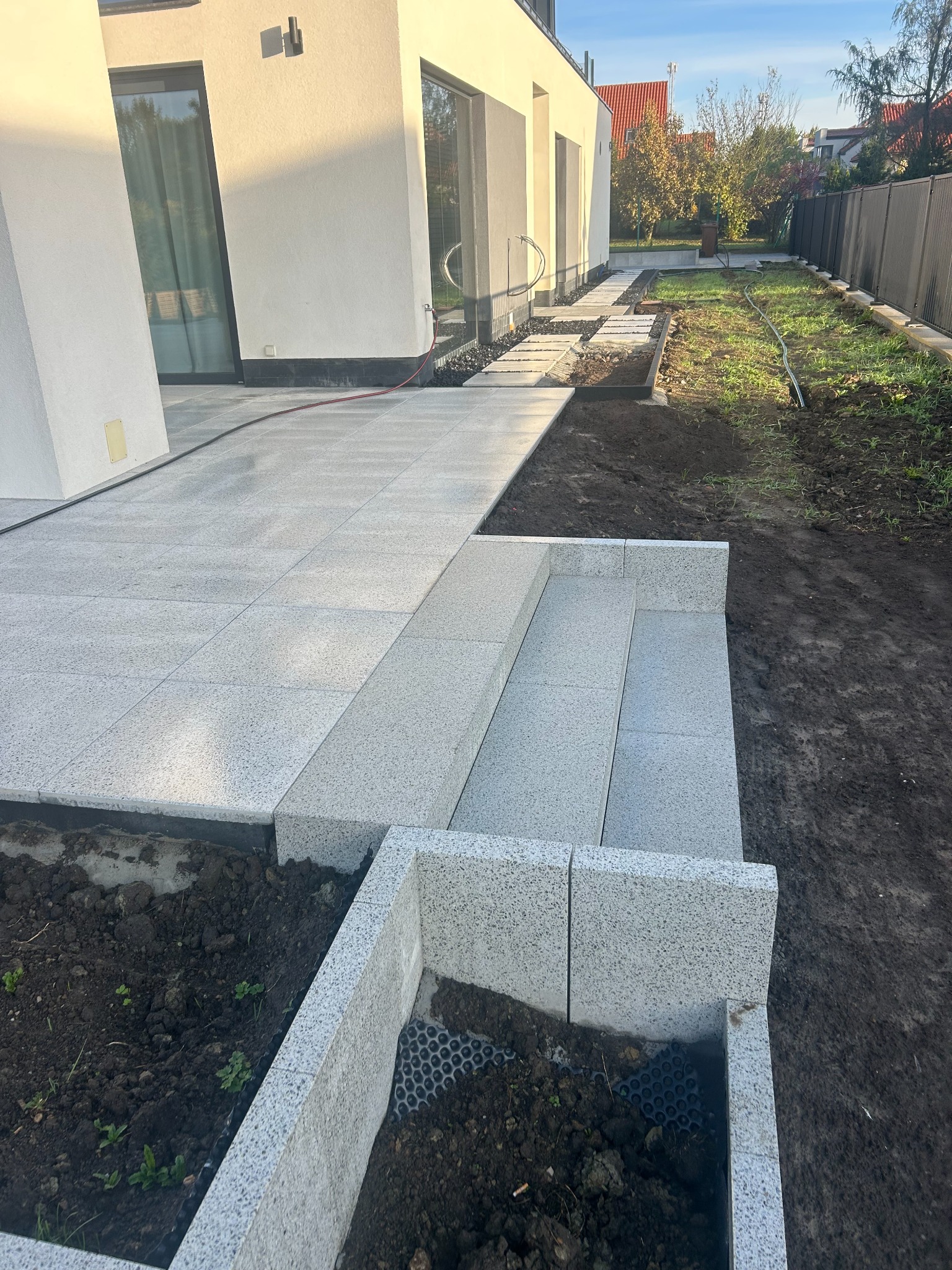 Jasnoszara kostka brukowa ułożona na ścieżce i schodach przy nowoczesnym domu. Widoczny fragment rabaty z obrzeżem z tej samej kostki. Tło: trawnik i ogrodzenie panelowe.