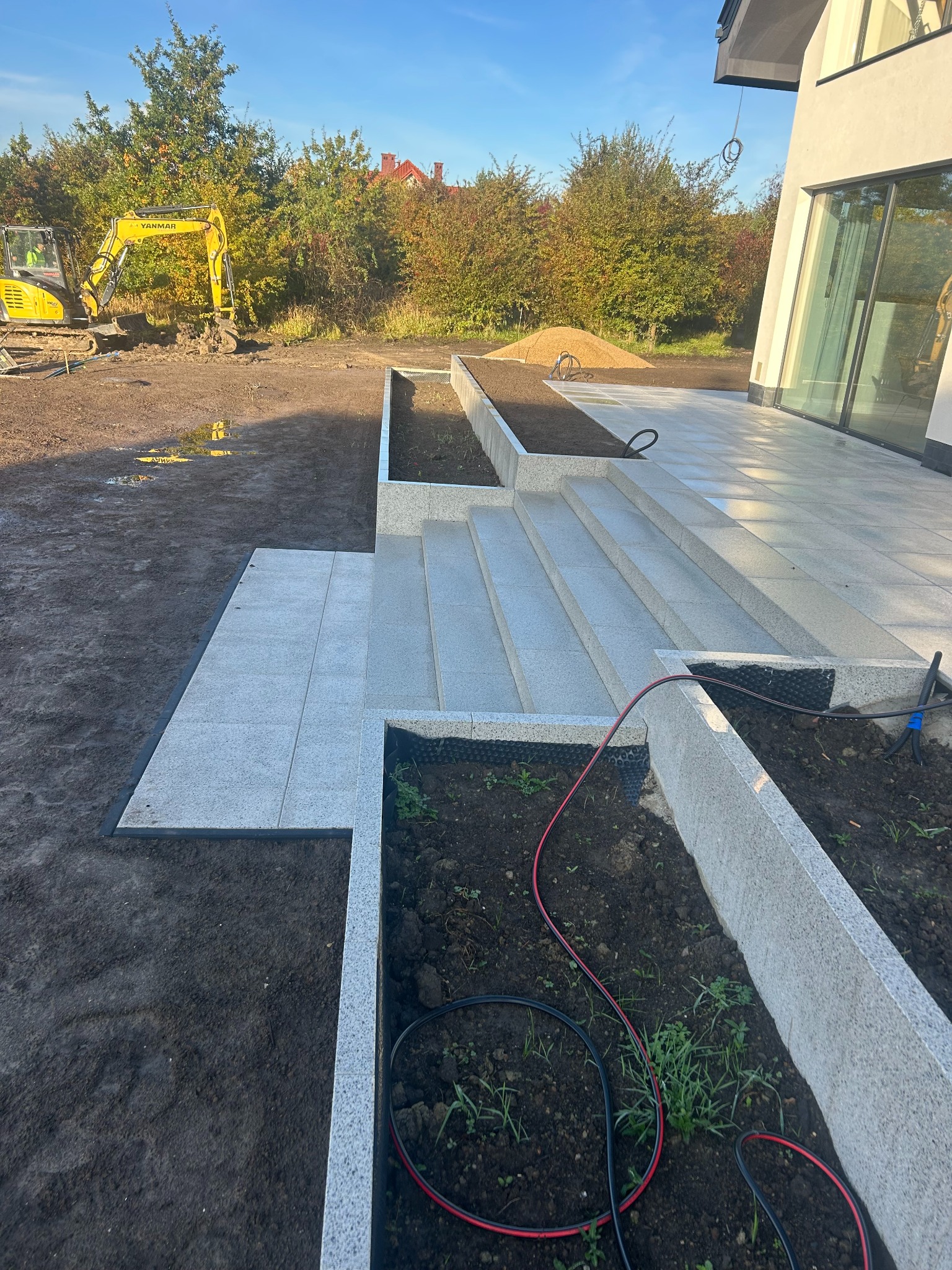 Granitowe schody i obrzeża rabat w ogrodzie, z żółtą koparką w tle. Widoczne przygotowane podłoże pod nasadzenia i świeżo ułożona nawierzchnia z jasnych płyt.