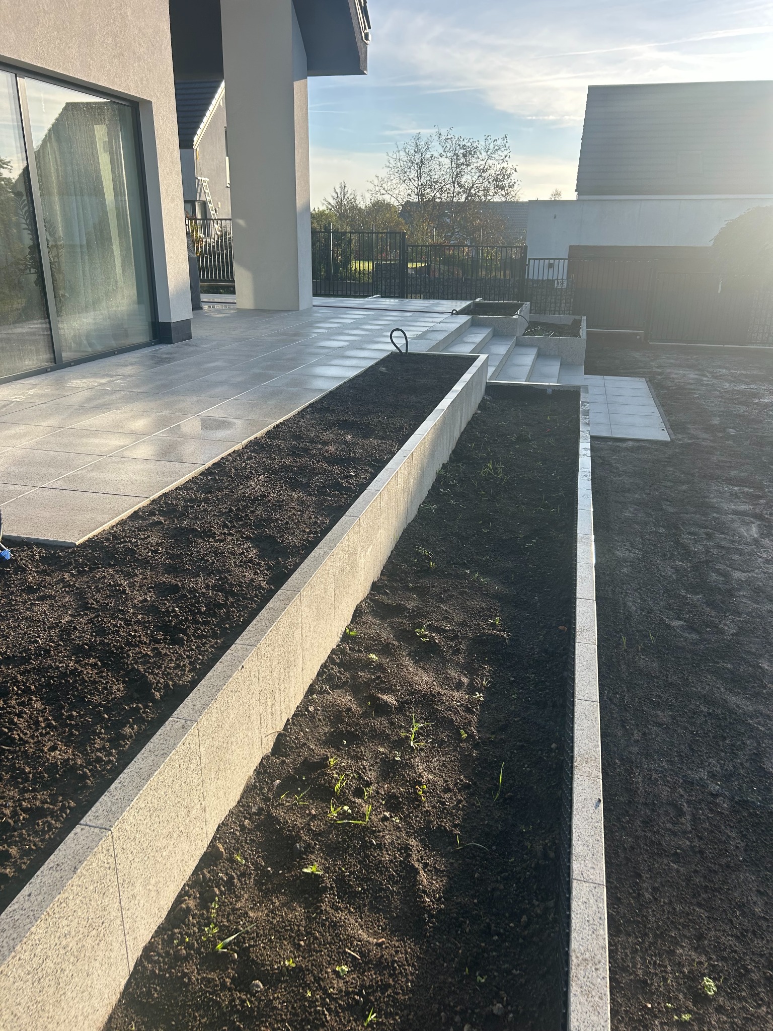 Nowoczesny taras z podwyższonymi rabatami wykończonymi jasną kostką, prowadzący do domu z dużym oknem przesuwnym. Minimalistyczny design, świeżo nasypana ziemia.
