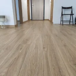 Beżowe panele podłogowe ułożone wzdłuż korytarza z widocznymi drzwiami wejściowymi z czarną ramą i brązowymi ościeżnicami, krzesło składane przy ścianie.