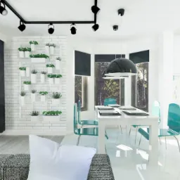 Jasna jadalnia z białym stołem, turkusowymi krzesłami, ścianą z cegły z roślinami w białych doniczkach i czarną lampą wiszącą. W tle widok na las przez okna.