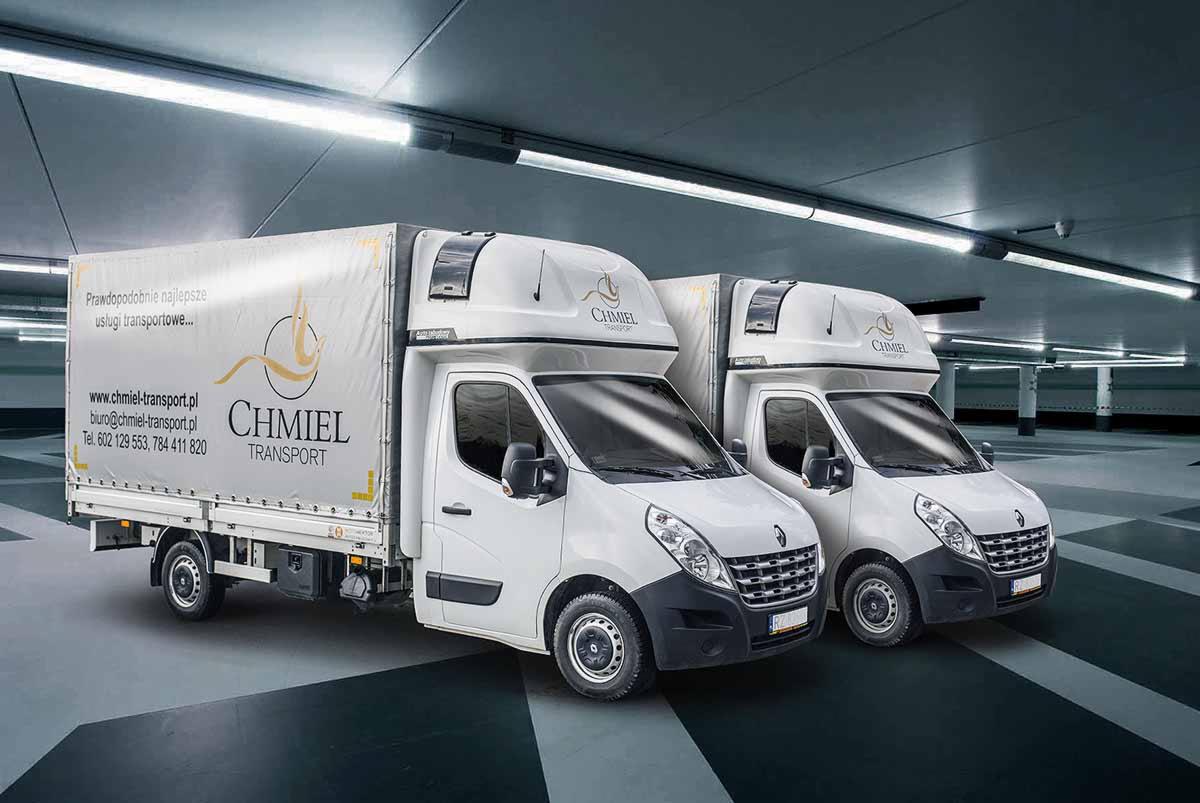 Dwa białe busy transportowe z logo firmy CHMIEL TRANSPORT na tle parkingu podziemnego, oświetlone jarzeniowym światłem.