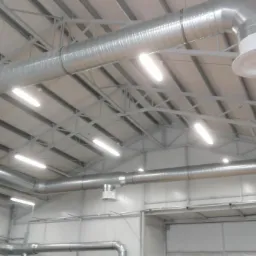 System kanałów wentylacyjnych z okrągłymi wylotami, zamontowany pod sufitem hali przemysłowej w Wydminach, oświetlony lampami jarzeniowymi.