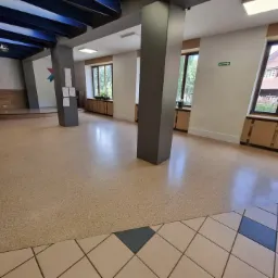 Przestronne, jasne wnętrze sali z terazzo na podłodze, szare filary, okna z roślinami doniczkowymi i drewnianymi szafkami pod parapetami, oraz małą sceną w tle. Widoczny fragment kafelkowej podłogi...