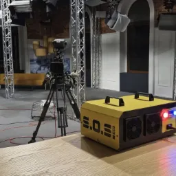 Żółty osuszacz powietrza S.O.S. 40 na drewnianym stole w studiu filmowym z kamerą na statywie i scenografią w tle.