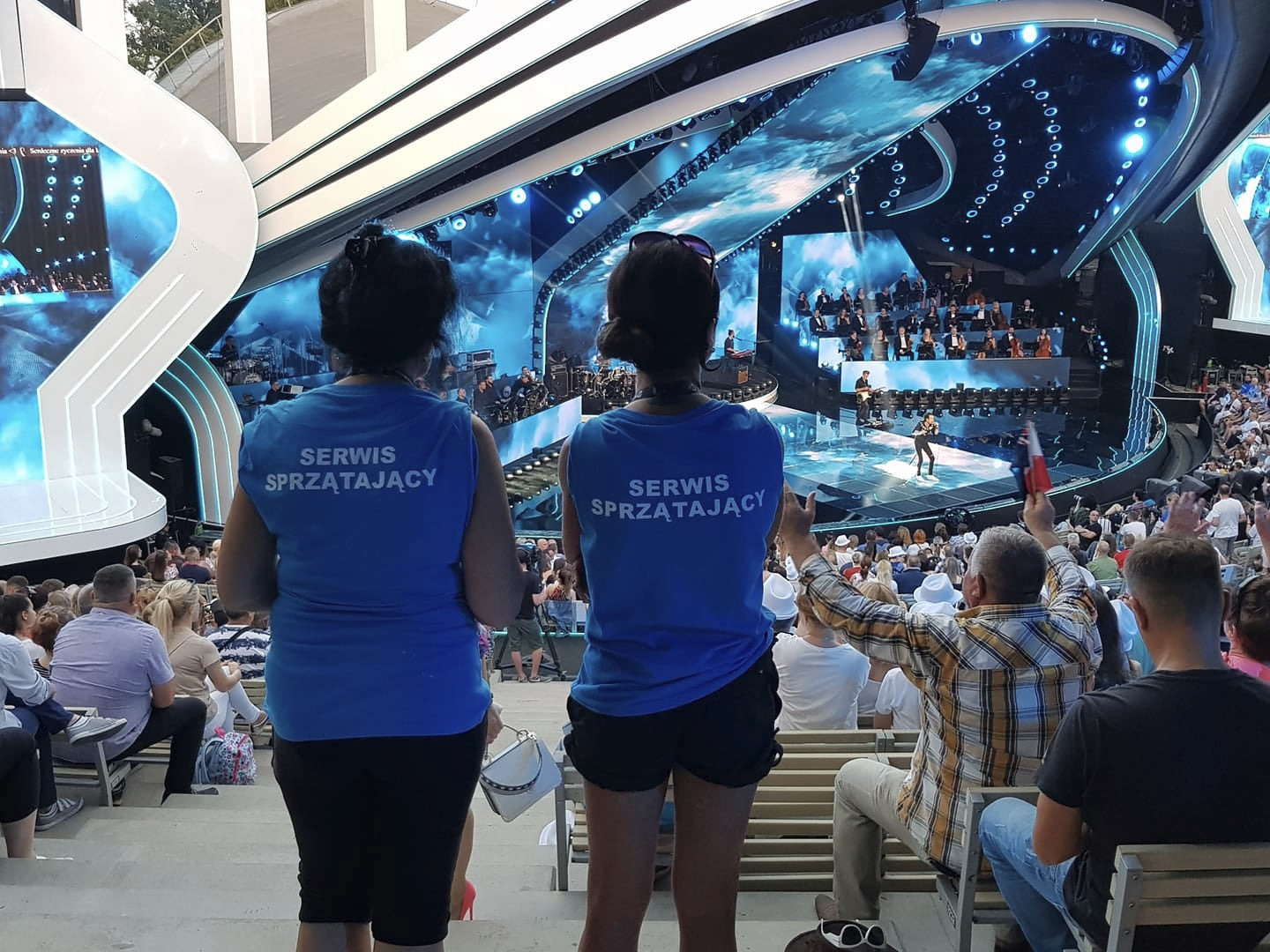 Dwie kobiety w niebieskich koszulkach z napisem 'Serwis Sprzątający' stoją na trybunach w Jełowej, obserwując scenę podczas wydarzenia kulturalnego.