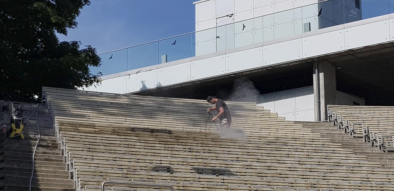 Mężczyzna w czapce i koszulce czyści trybuny stadionu za pomocą myjki ciśnieniowej, widoczne smugi brudu i strumień wody.