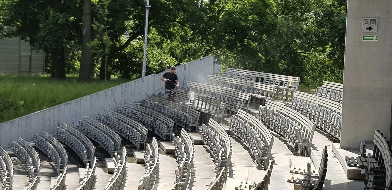 Mężczyzna w szortach i czarnej koszulce czyści pod wysokim ciśnieniem rzędy krzeseł na betonowym amfiteatrze w słoneczny dzień.