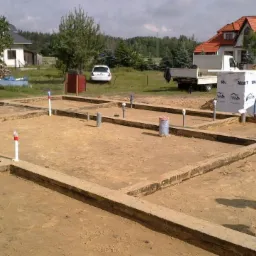 Fundamenty domu jednorodzinnego w trakcie budowy, widoczne szalunki i wystające rury instalacyjne. W tle materiały budowlane i krajobraz wiejski.