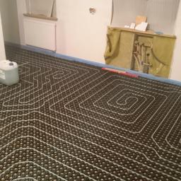 Revo Instal - kompleksowe wykonawstwo instalacyjne - Ułożona instalacja ogrzewania podłogowego z widocznymi rurami grzewczymi, przed wylaniem posadzki. Białe ściany i okno w tle. Kanister z płynem obok.