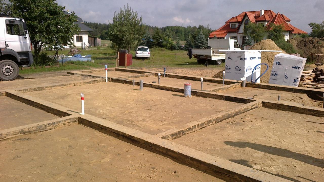 Fundamenty domu jednorodzinnego w trakcie budowy, widoczne szalunki i wystające rury instalacyjne. W tle materiały budowlane i krajobraz wiejski.