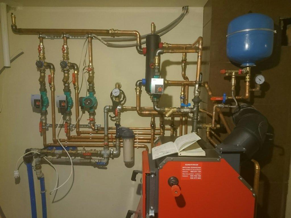 Złożona instalacja grzewcza z licznymi rurami miedzianymi, zaworami i pompami, zamontowana na ścianie w kotłowni. Na pierwszym planie czerwony piec z otwartą książką.