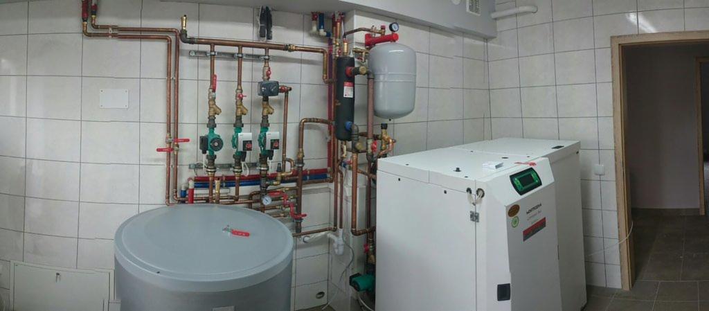 Złożona instalacja grzewcza z kotłem i zasobnikiem w pomieszczeniu wyłożonym kafelkami. Widoczne liczne rury miedziane, zawory i pompy obiegowe.