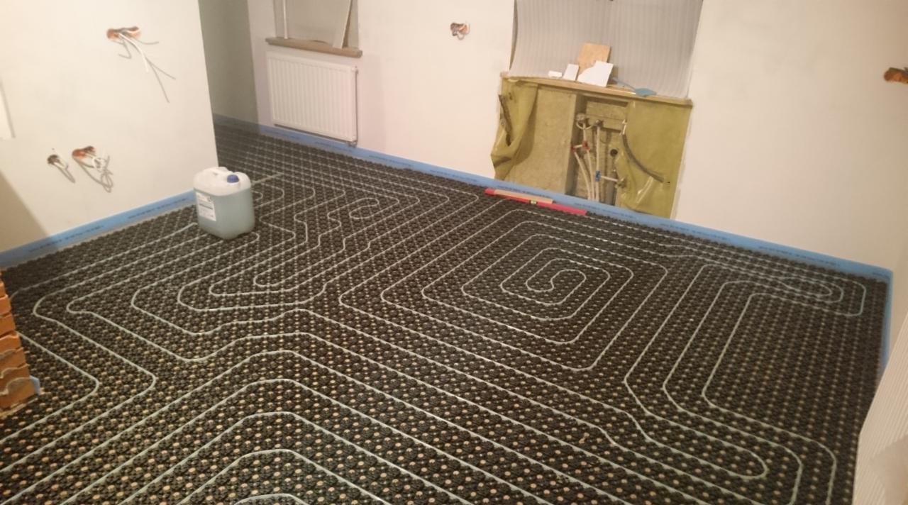 Ułożona instalacja ogrzewania podłogowego z widocznymi rurami grzewczymi, przed wylaniem posadzki. Białe ściany i okno w tle. Kanister z płynem obok.