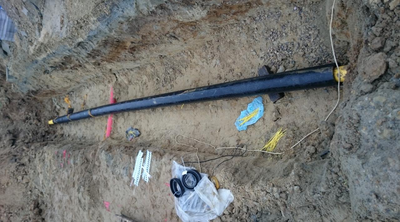 Czarna rura w wykopie ziemnym, zabezpieczona żółtymi zaślepkami. Widoczne akcesoria instalacyjne i oznaczenia geodezyjne. Instalacja podziemna w trakcie realizacji.