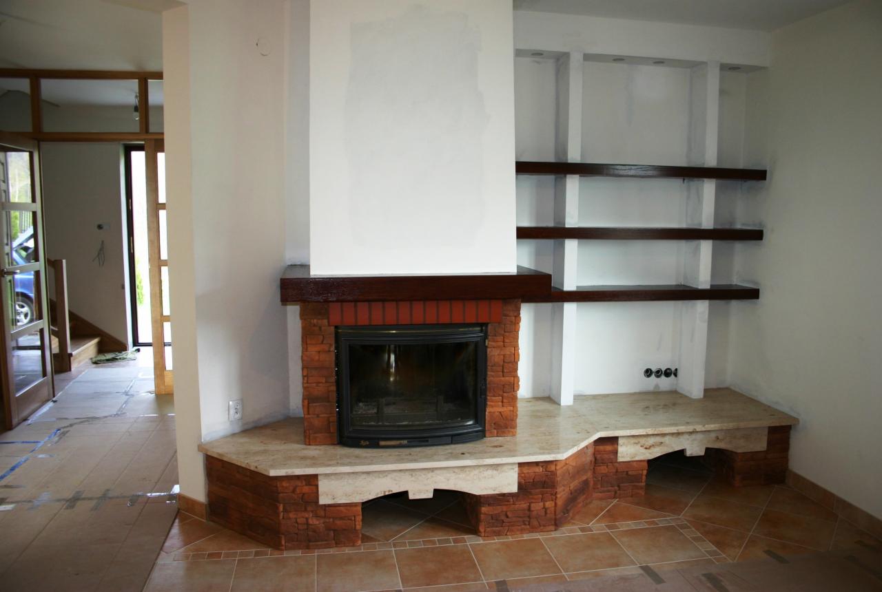 Murowany kominek z półkami wnękowymi w jasnym salonie, widok z boku, płytki na podłodze częściowo zakryte kartonem zabezpieczającym.