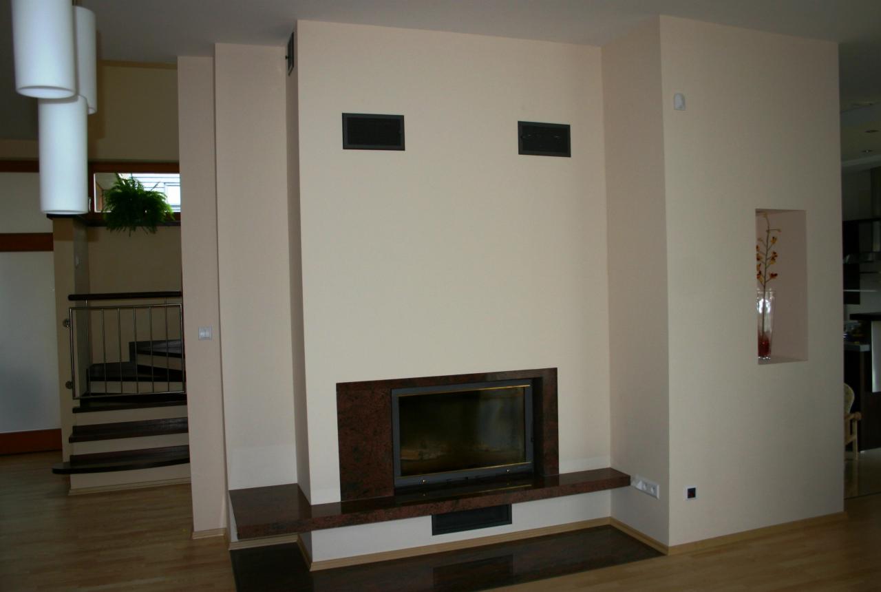 Nowoczesny kominek z obudową z ciemnego granitu, wbudowany w ścianę w jasnym salonie. Widoczna dekoracyjna wnęka ścienna z wazonem. Schody w tle.