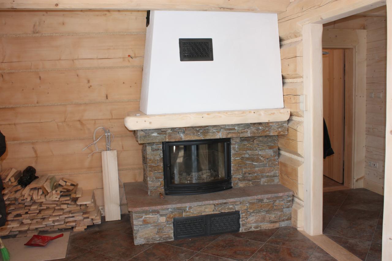 Kominek z obudową z kamienia naturalnego w drewnianym wnętrzu. Widoczna część paleniska i kratka wentylacyjna. Styl rustykalny, ciepłe oświetlenie.