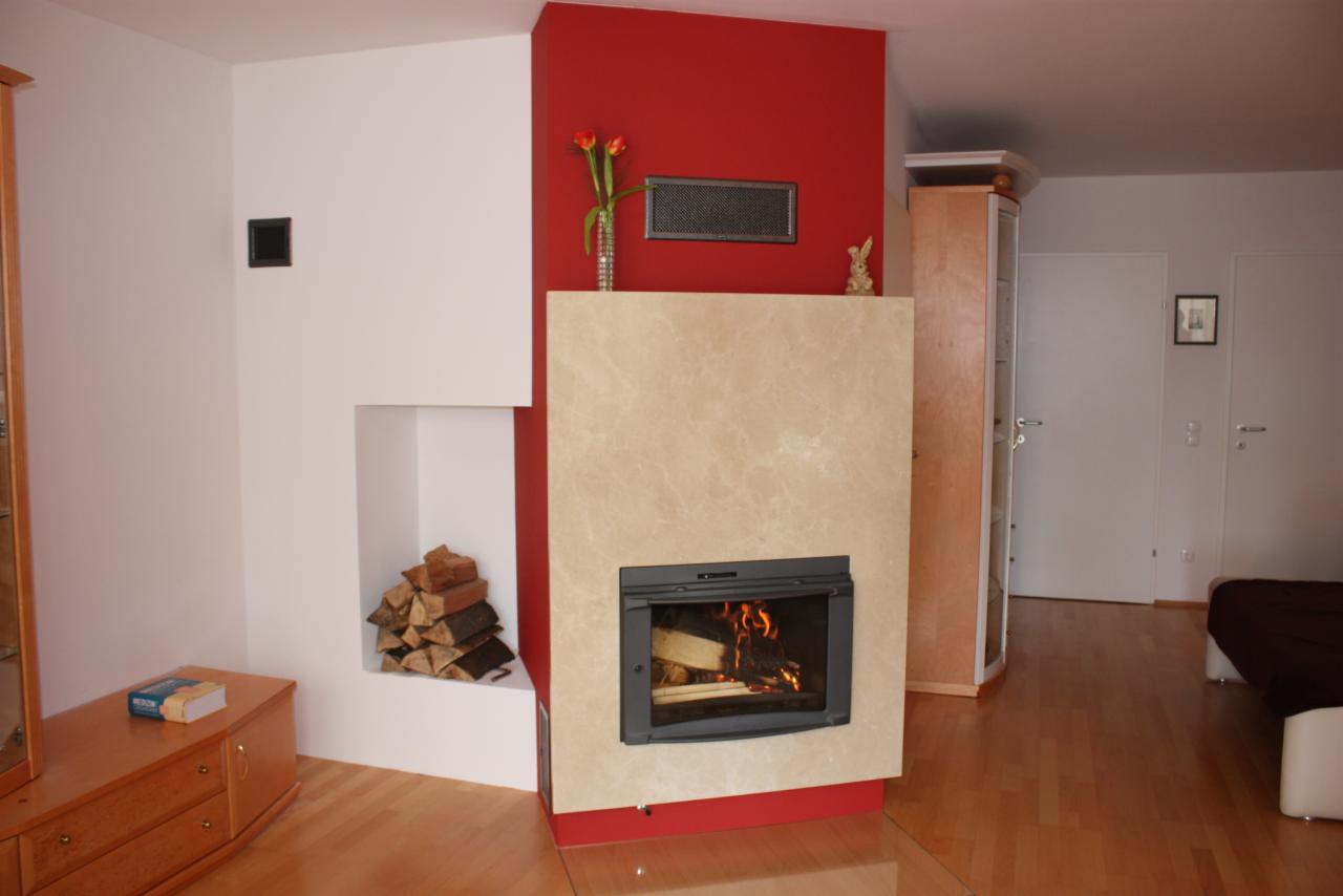 Nowoczesny kominek z marmurową obudową i paleniskiem, z zapasem drewna obok, na tle czerwonej ściany. Uwagę przykuwa detal: figurka zajączka na kominku.