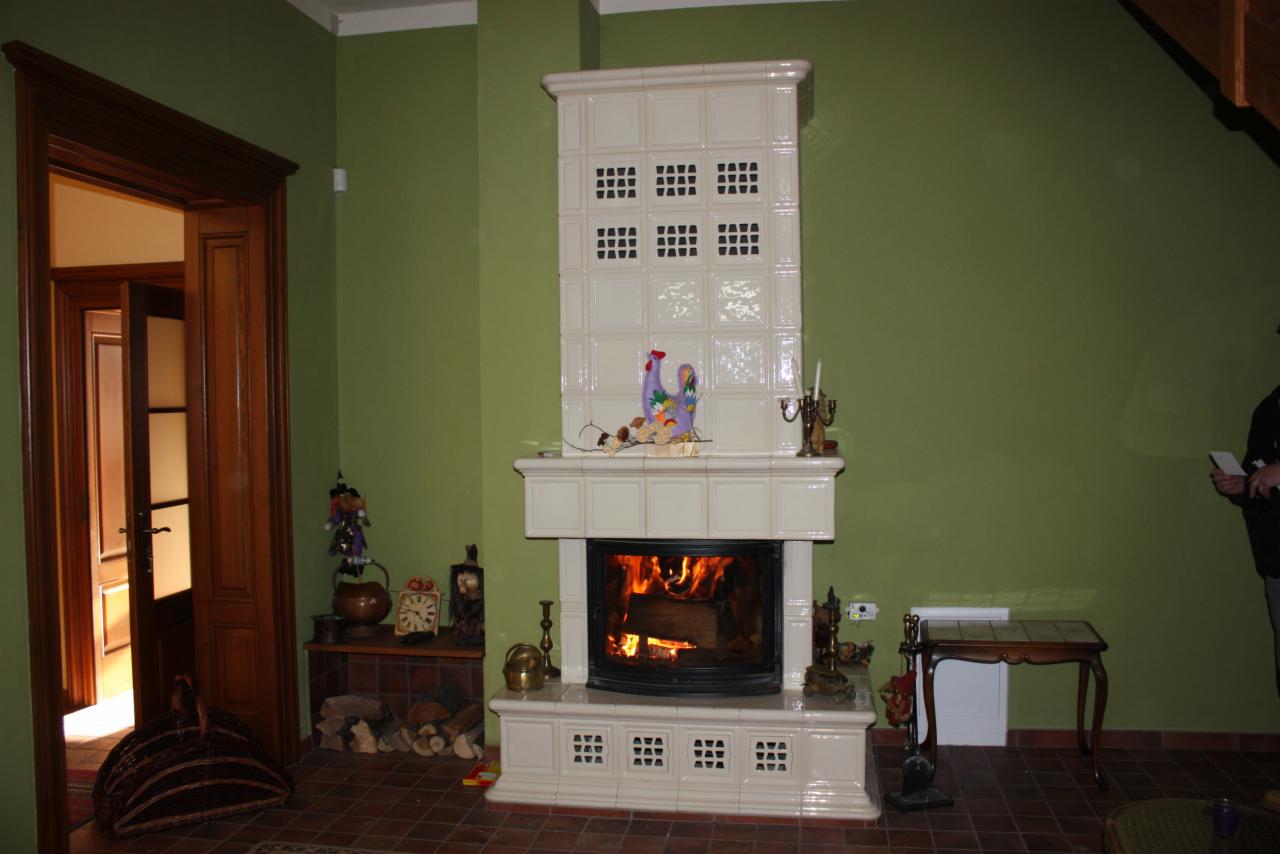 Biały, kaflowy kominek z paleniskiem i dekoracjami w oliwkowym salonie. Widoczny fragment drewnianych drzwi i dekoracje obok kominka. Płomienie w palenisku.