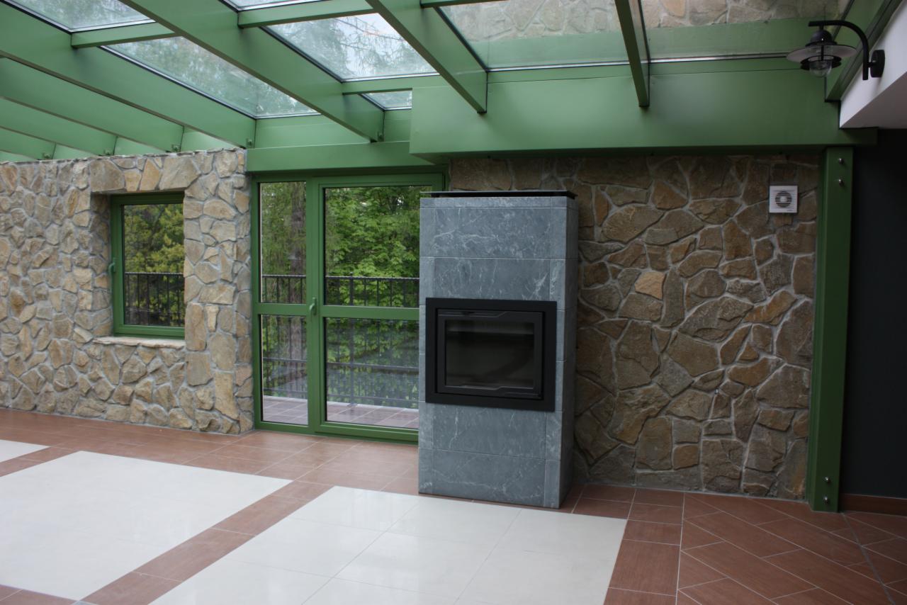 Murowany kominek z szarego kamienia w przeszklonym pomieszczeniu z kamienną ścianą i zielonymi ramami okiennymi. Widok na zieleń za oknem.