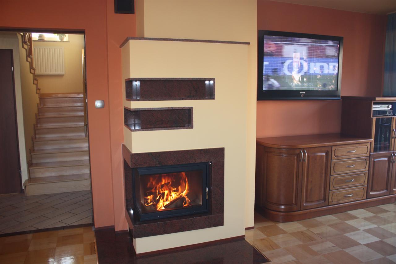 Nowoczesny kominek z obudową z ciemnego granitu, z płomieniami widocznymi przez szybę, w salonie z telewizorem i drewnianymi meblami. W tle schody.