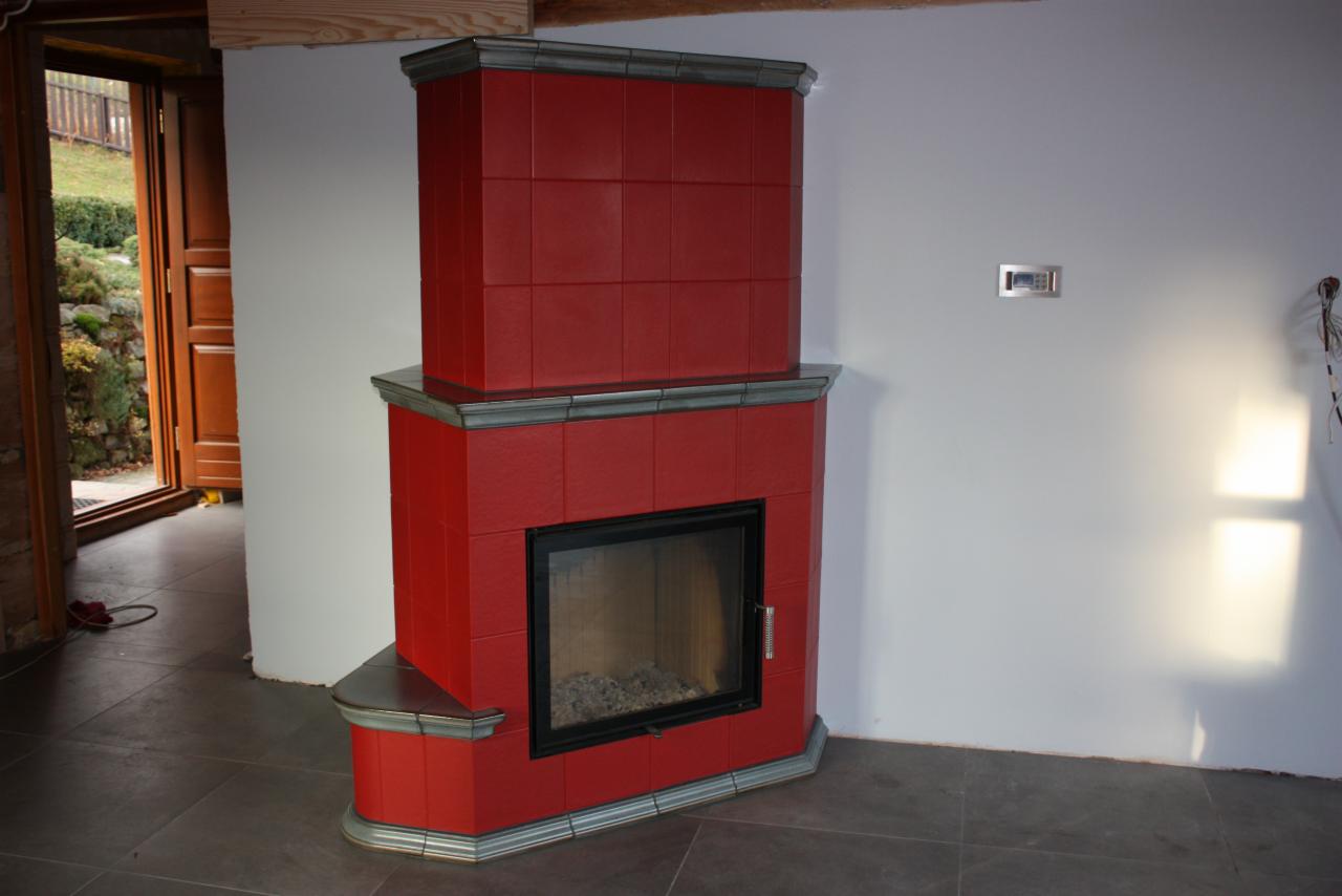 Czerwony, kaflowy kominek z czarną ramą, w rogu pokoju z szarą podłogą i otwartymi drzwiami na zewnątrz. Widoczny fragment drewnianej belki stropowej.