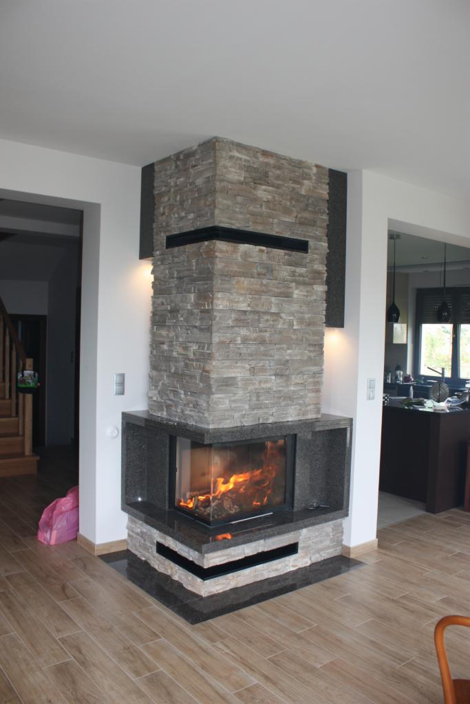 Narożny kominek z obudową z kamienia i granitu, z paleniskiem wypełnionym płomieniami, w nowoczesnym salonie z drewnianą podłogą i widokiem na kuchnię.