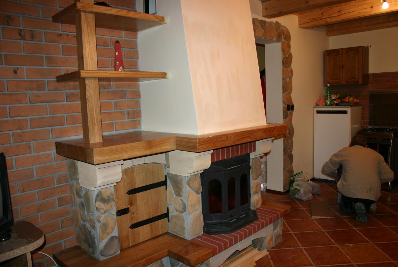 Rustykalny kominek z kamienia i drewna z półkami, ceglana ściana w tle i osoba w kuchni. Unikalna zabudowa kominkowa w trakcie wykańczania.